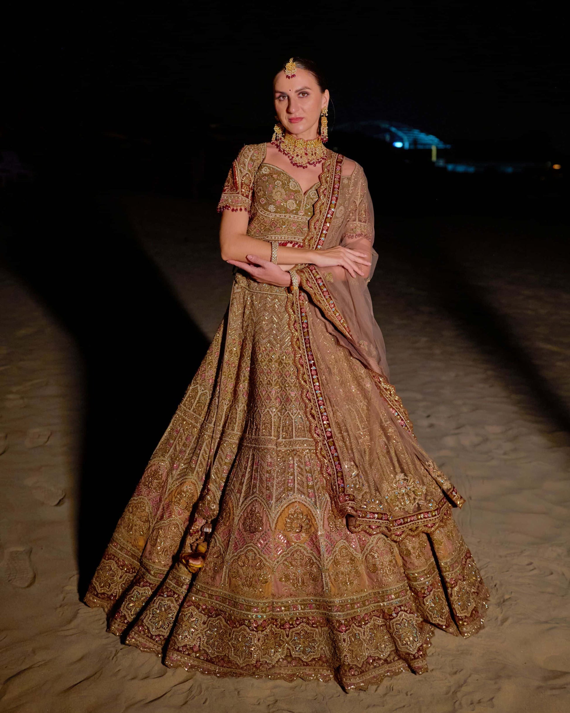Woman in an ornate traditional dress standing on a sandy surface at night.