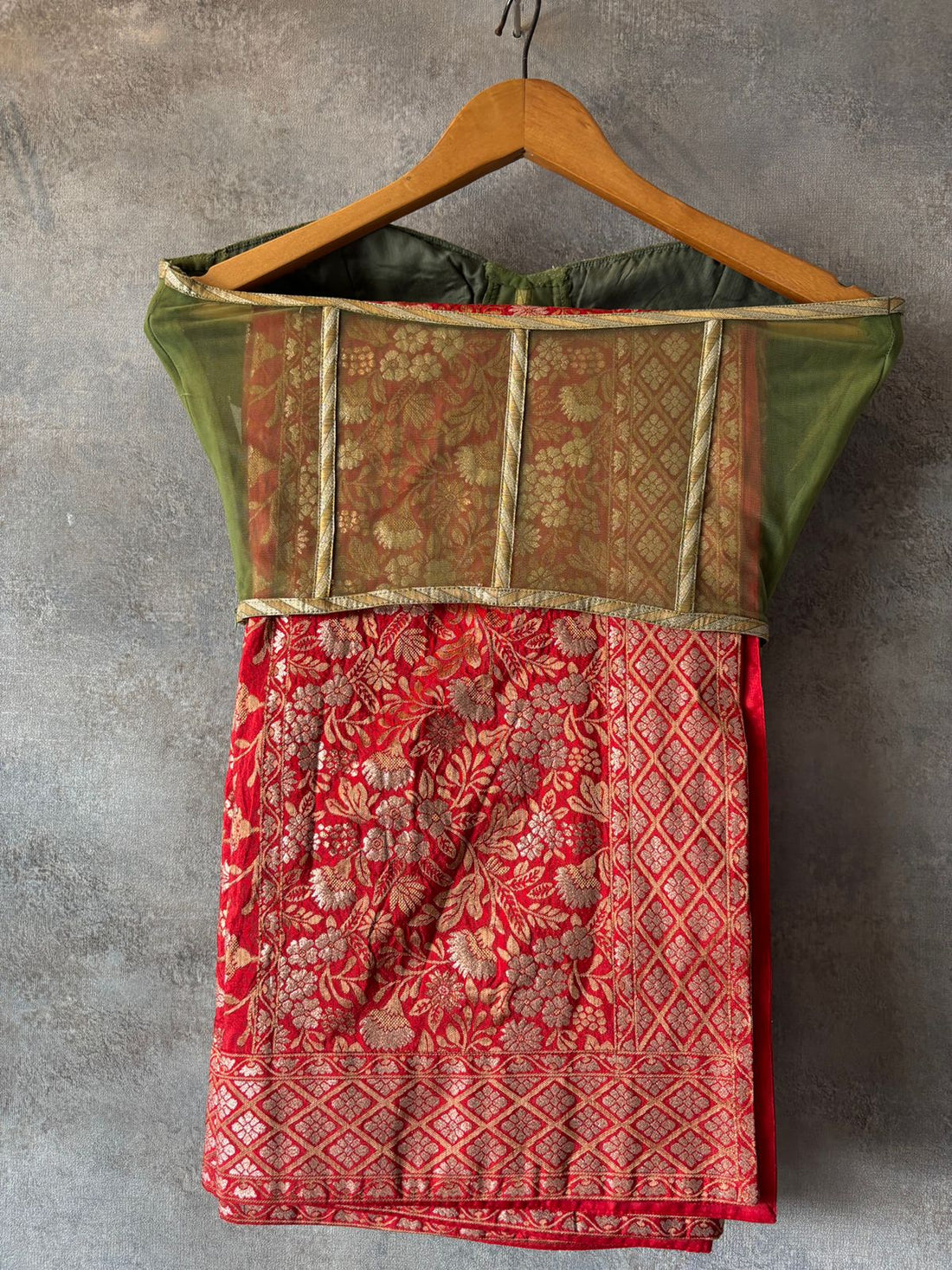 Traditional outfit with a red blouse and green and white patterned skirt on hangers against a gray wall.